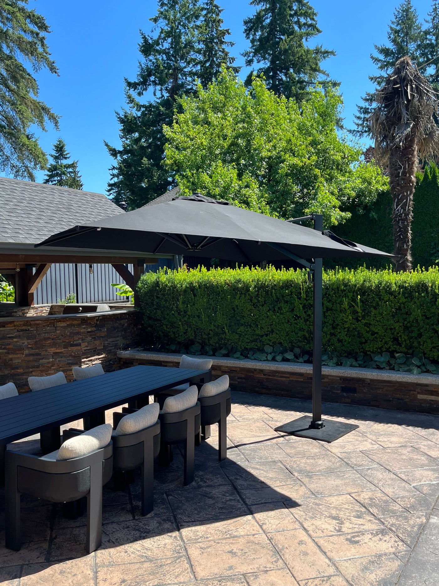 RM Sun Umbrella 10x10 cantilever, rotatable, providing shade over a patio dining table in a lush garden setting.
