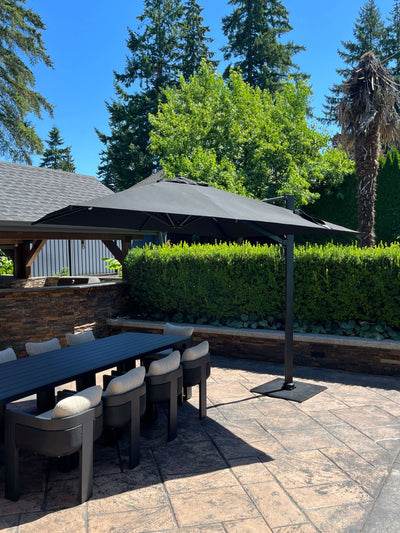 RM Sun Umbrella 10x10 cantilever, rotatable, providing shade over a patio dining table in a lush garden setting.